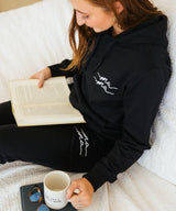 Person wearing a black hoodie with white designs, sitting on a couch holding a book and mug.