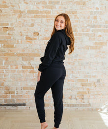 A woman in a black outfit stands against a brick wall.