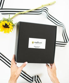 Black box with a brand logo held by hands on a white background with sunflower and ribbons.