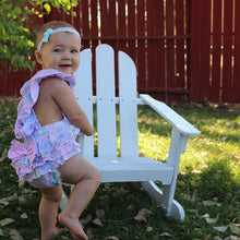 Baby in a colorful outfit sitting on a white chair outdoors.