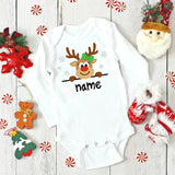 White baby onesie with a reindeer design and 'name' text, surrounded by Christmas decorations on a wooden surface.