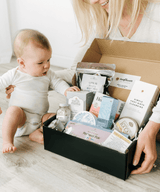 Woman and baby with a box of products on a light wooden floor.