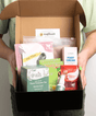 Open box containing various products with visible branding, held by a person.