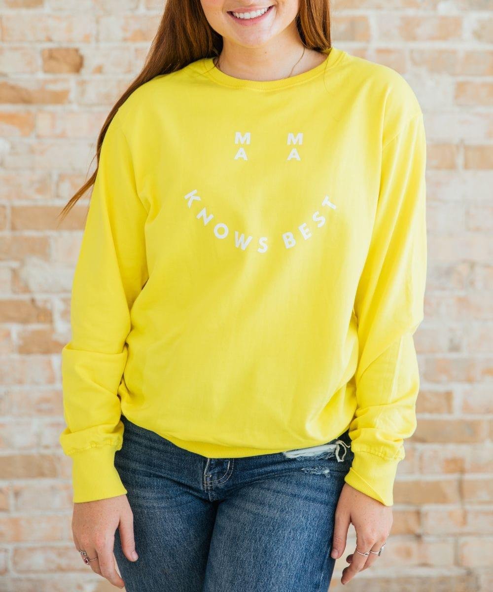 Person wearing a yellow sweatshirt with 'MAMA KNOWS BEST' text against a brick wall.
