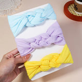 Three colorful knotted headbands (blue, purple, yellow) on a white surface with a hand holding one of them.