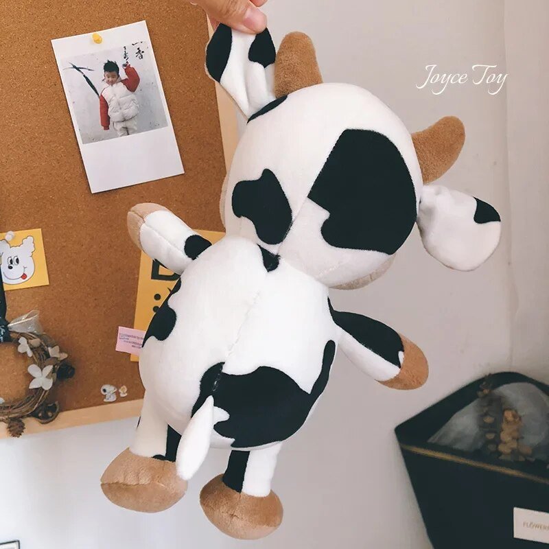 Plush cow toy held by a hand against a background with a cork board and photos.