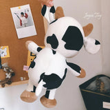 Plush cow toy held by a hand against a background with a cork board and photos.