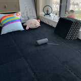 Dark quilted blanket on a bed with a pink plush toy, rainbow pillow, and black textured cube.