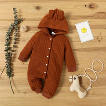 Brown baby onesie with buttons on a wooden surface, next to a toy and decorative elements.