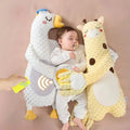 Baby lying between a white goose pillow and a yellow giraffe pillow while a hand holds a remote