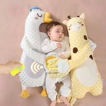 Baby lying between a white goose pillow and a yellow giraffe pillow while a hand holds a remote