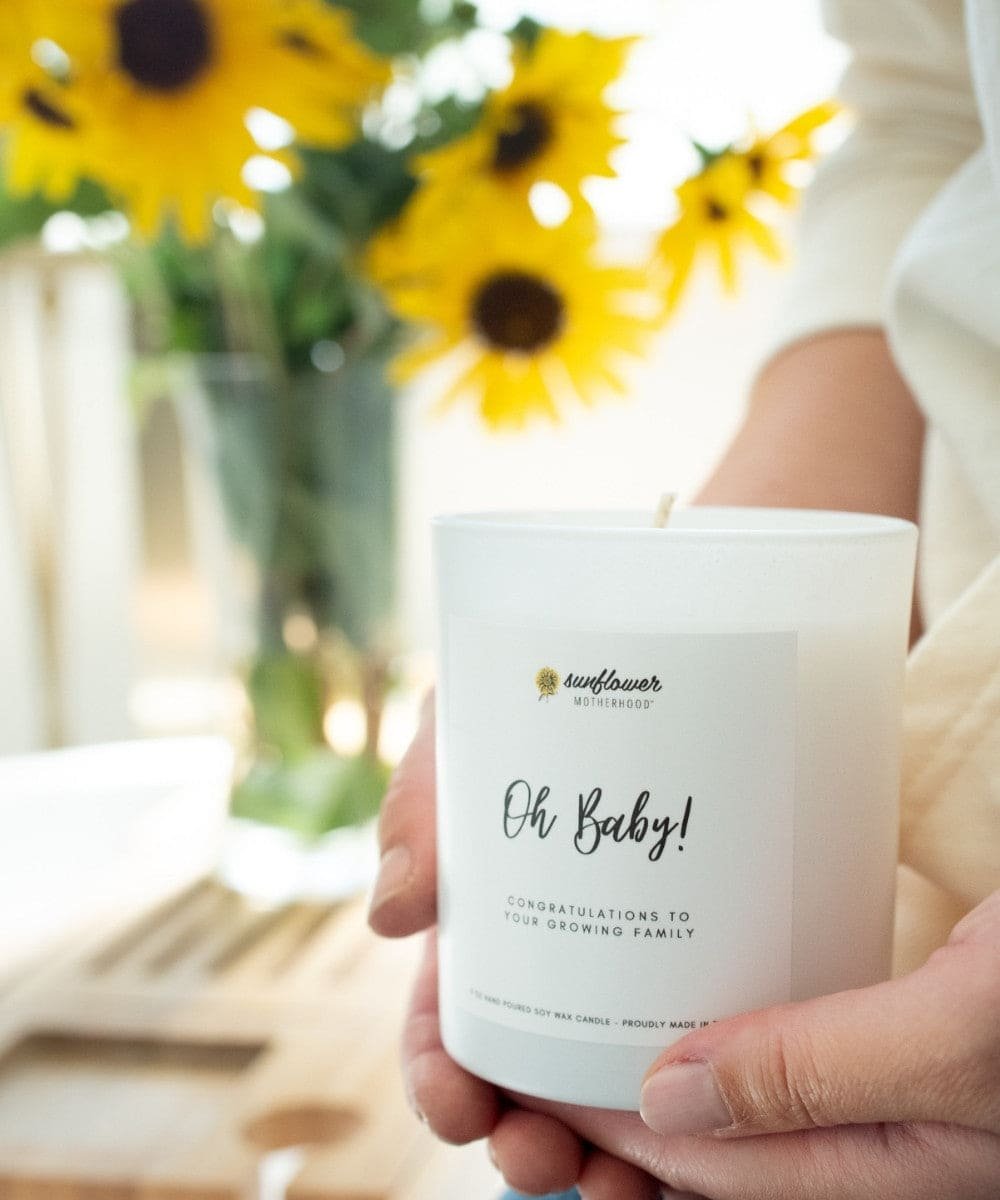 Person holding a 'Oh Baby!' candle with sunflowers in the background