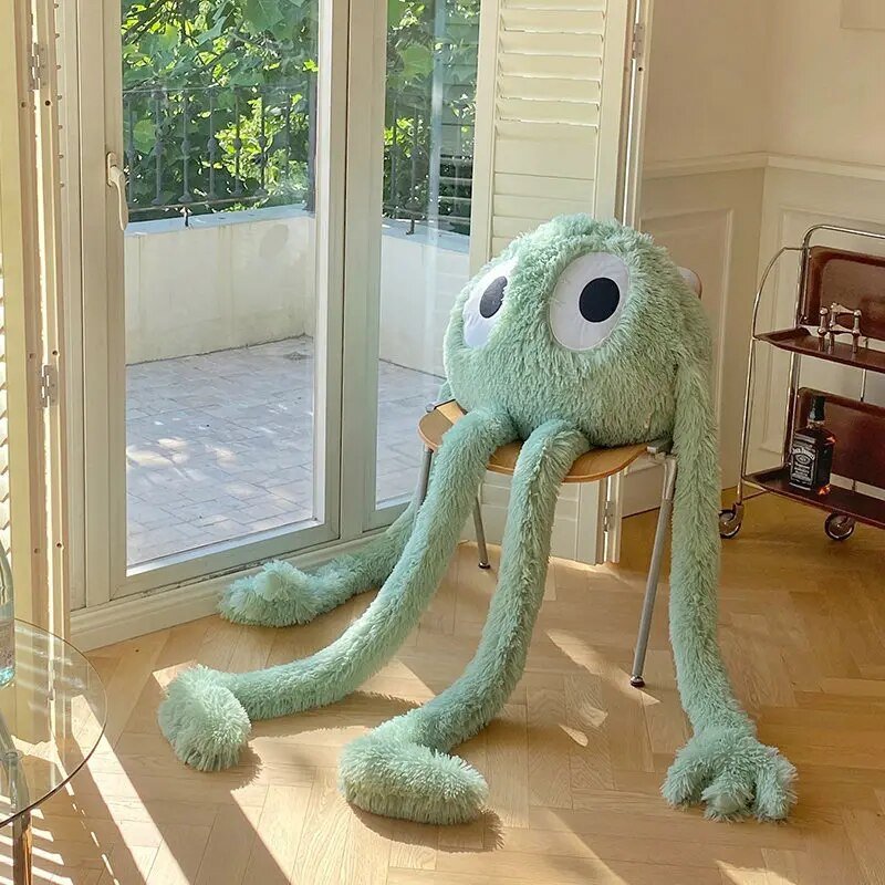 Super size 175cm green alien plush monster with long limbs seated in a bright sunlit room.