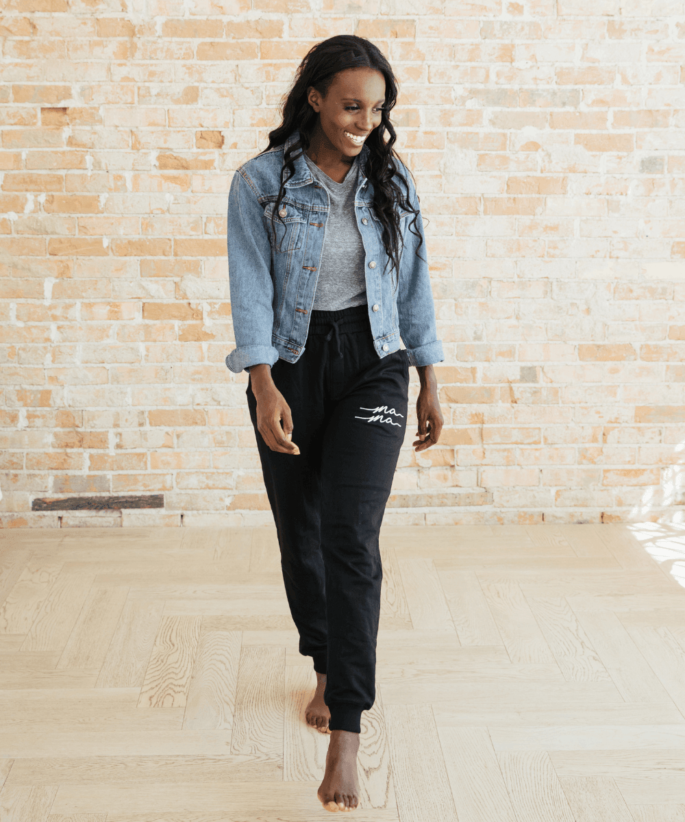 Woman wearing a denim jacket and black pants with white text against a brick wall.