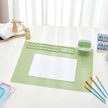 Green art desk mat with paper, paintbrushes, and a cup on a white surface.