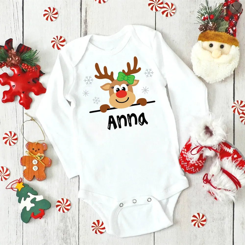 White baby onesie with a reindeer design and 'Anna' text, surrounded by Christmas decorations on a wooden surface.